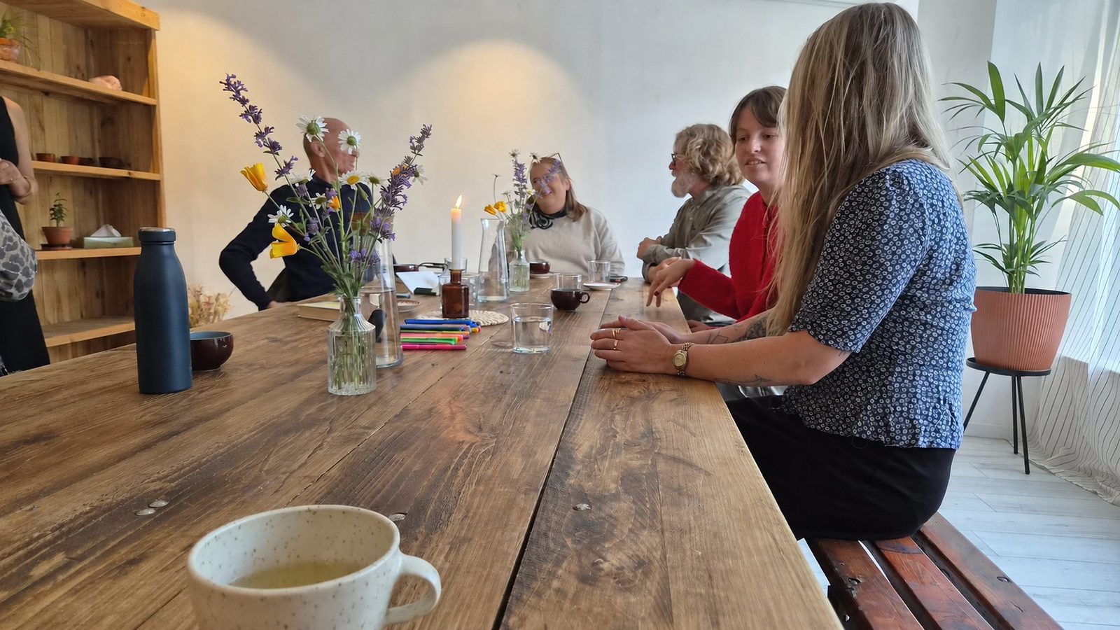 A refloria workshop in progress: a small group talking around a long wooden table with wildflowers, coloured pens, tea and a lit candle.